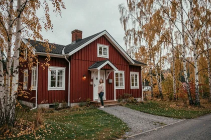 Femme devant une maison traditionnelle suédoise en bois rouge entourée de bouleaux en automne, illustrant l'achat immobilier en Suède