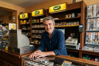 Gérant d'un bureau de tabac dans l'Ain debout derrière son comptoir avec une expression professionnelle et sereine
